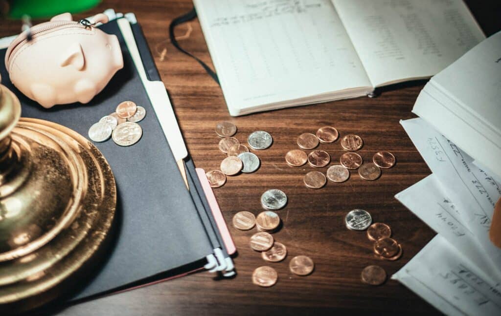 Imagem de moedas, folhas de papel, caderno, pasta e um porquinho de dinheiro em uma superfície de madeira iluminada.
