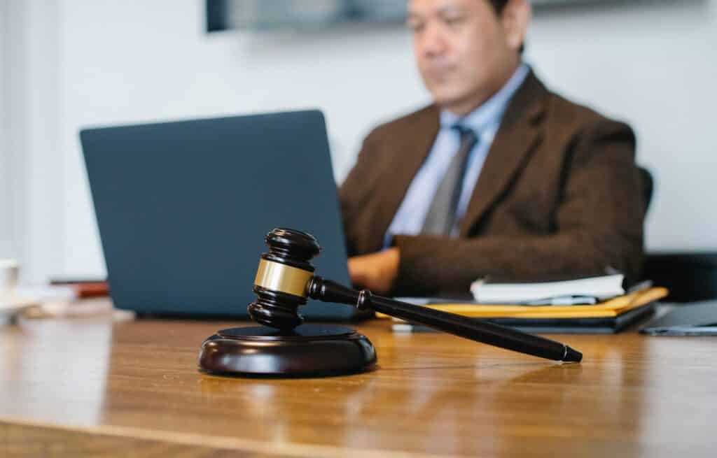 Imagem com foco em um martelo utilizado por juízes em julgamentos nos tribunais e, ao fundo, uma pessoa mexendo em um notebook em desfoque.