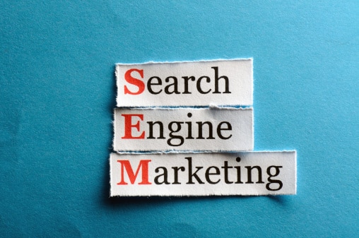Fotografia de três recortes com as palavras Search, Engine e Marketing, respectivamente. A junção forma o termo SEM. O fundo é um papel azul, levemente amassado.