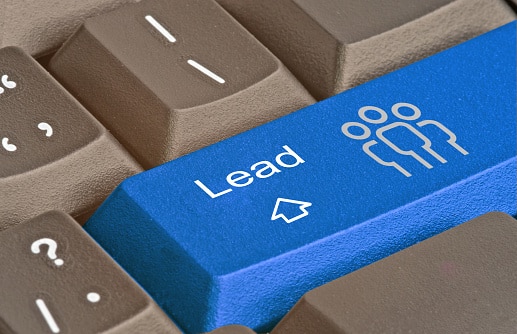 Fotografia de um teclado cinza e que possui um botão azul com a palavra "lead", o qual também é composto por uma seta na direção do termo e um ícone de "pessoas", todos na cor branca.